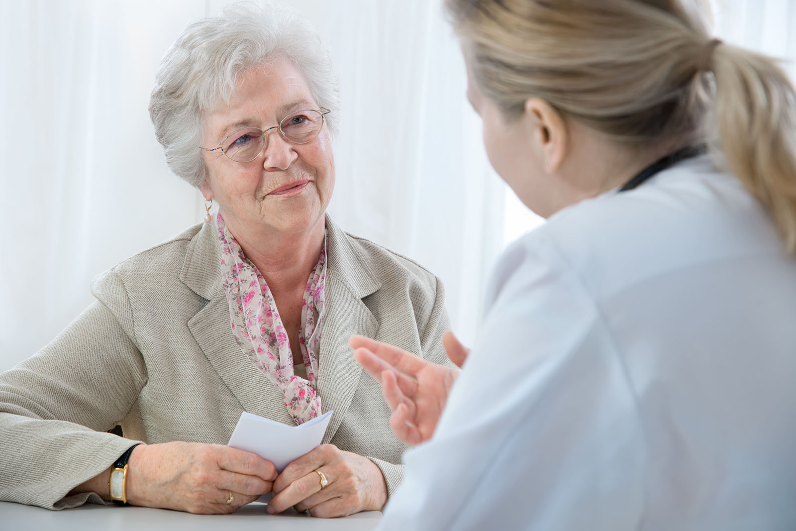 Eine ältere Patientin und eine Ärztin unterhalten sich sitzend am Tisch.