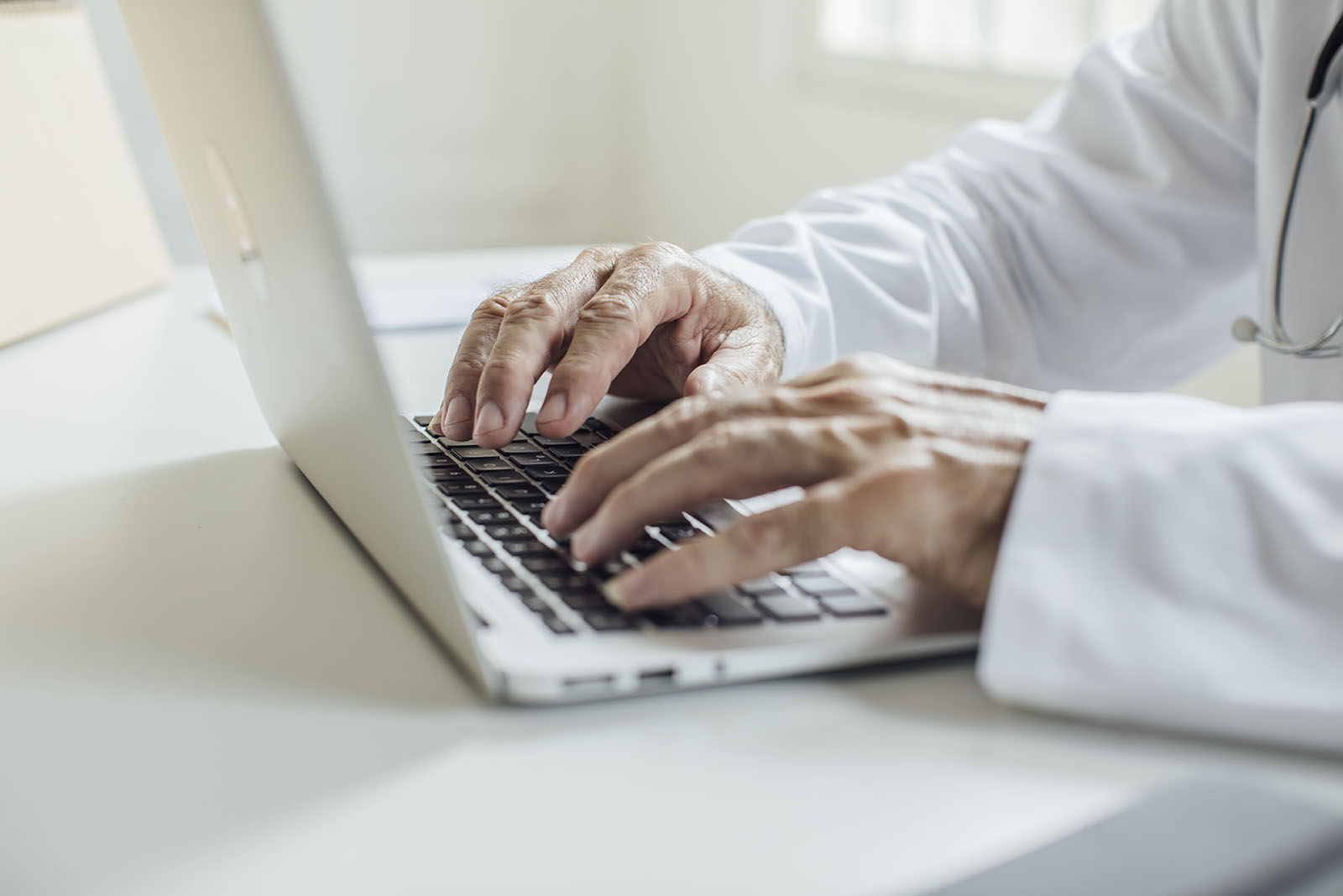 Ein weißer Laptop steht auf einem weißen Tisch. Beide Hände eines Arztes tippen auf der Tastatur des Laptops.