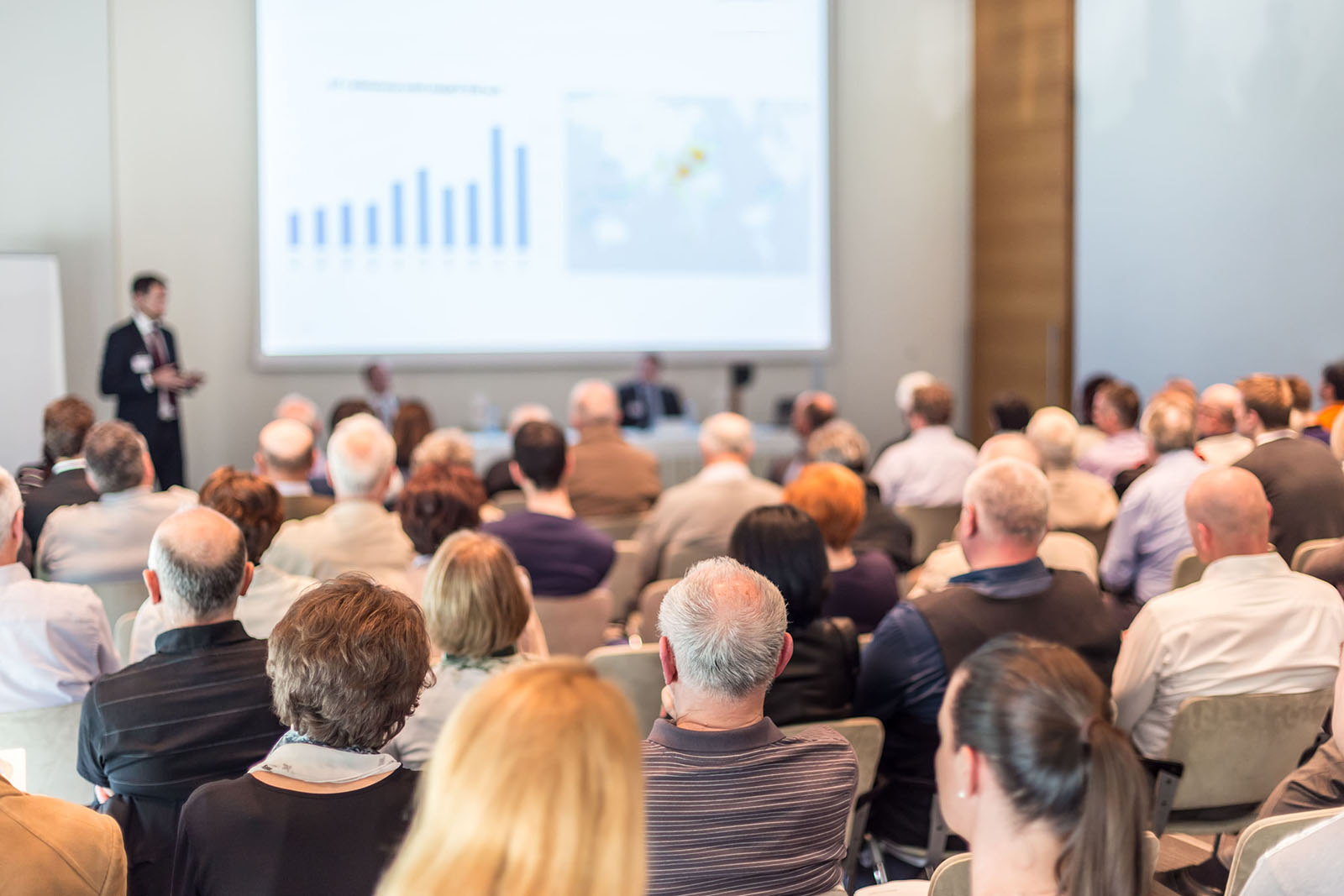 Viele Menschen sitzen in einem Saal und verfolgen einen Vortrag. Auf der Leinwand sind blaue Grafiken zu erkennen, links daneben steht der Vortragende.