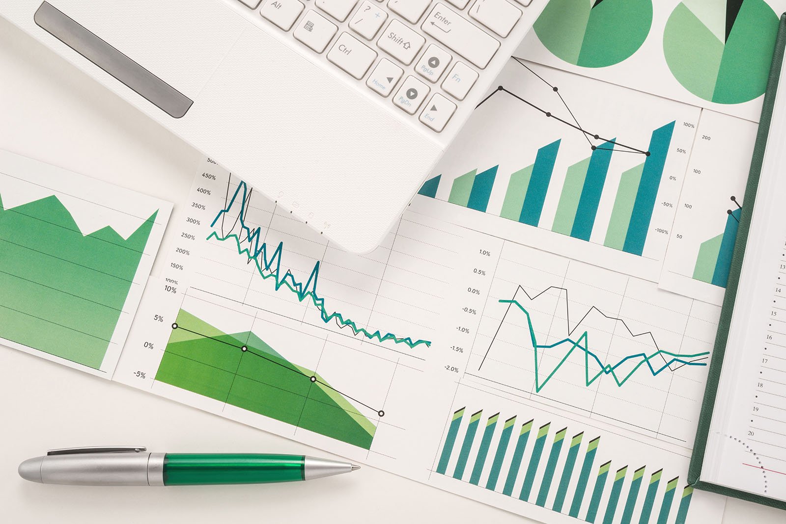 Ausschnitt eines weißen Laptops, der auf weißen Blättern mit verschiedenen grünen, grauen und blauen Diagrammtypen liegt. Daneben liegt ein silber-grüner Kugelschreiber.